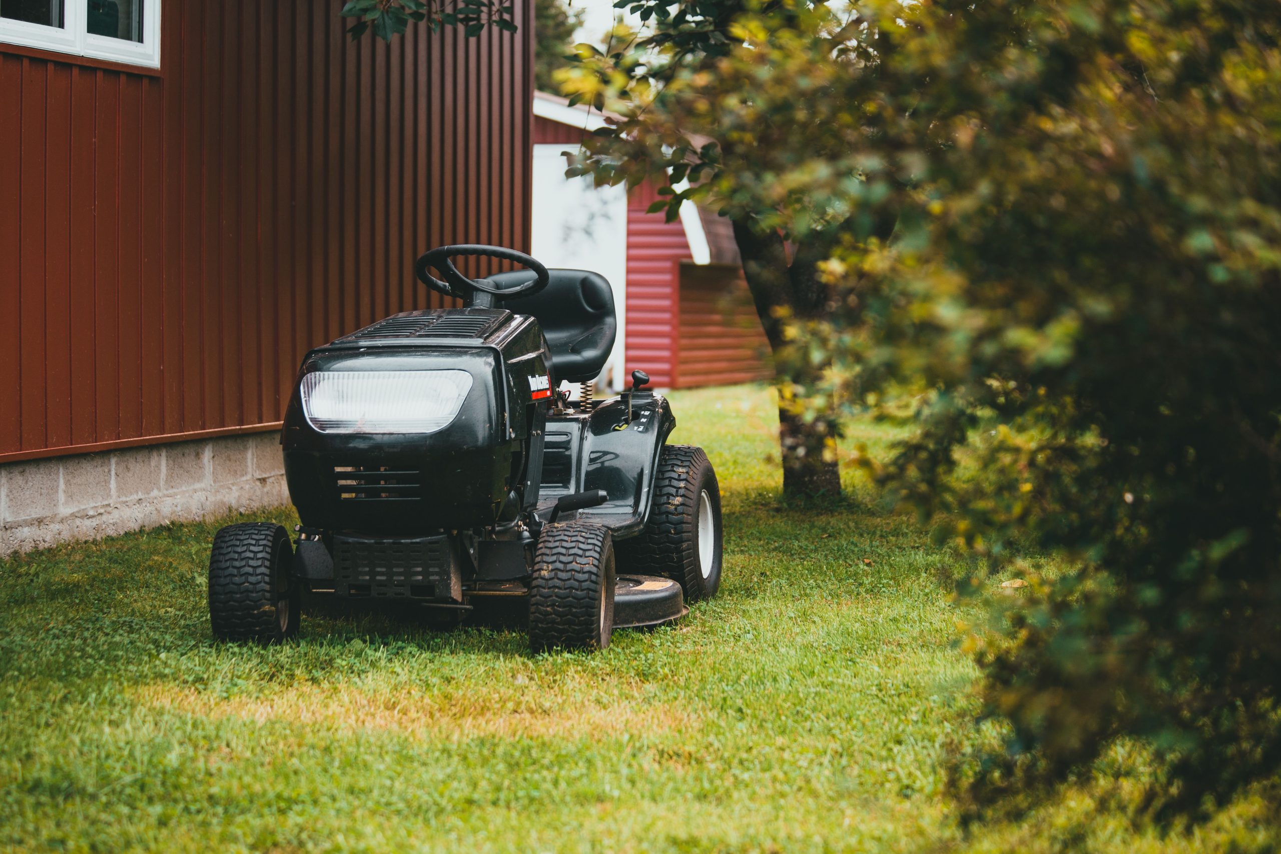A black ride-on lawnmower parked on green grass near a red barn during summer.